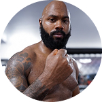 A bald, bearded man with tattoos on his shoulder and arm clenches his fist.