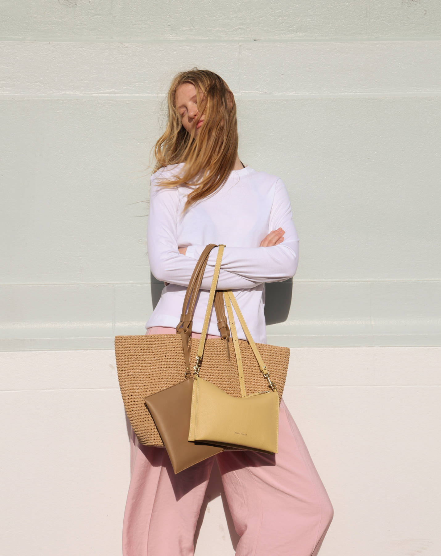 Person with straw bag and pink pants, standing against a light wall.