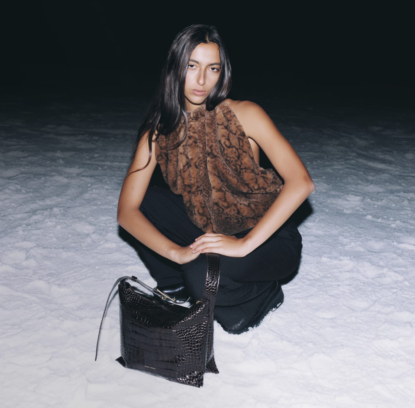 Person squatting on snow at night, wearing a brown top, with a black bag.