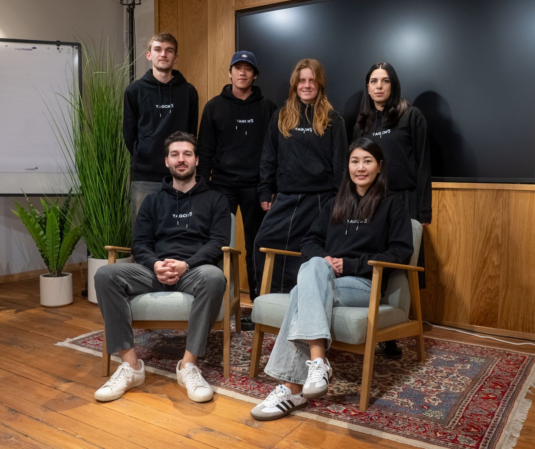 Group of six people in black hoodies posed in a room with plants and a screen.
