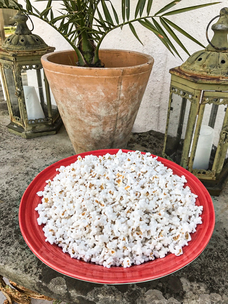 A red plate of popcorn sits on a stone surface in front of a potted plant and two lanterns.