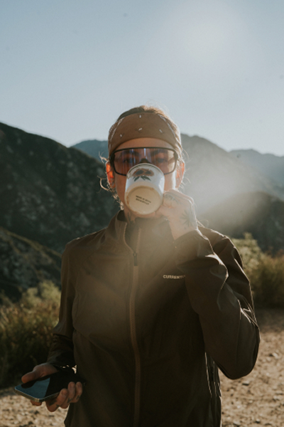 Person drinking from a mug outdoors, holding a phone, with mountains in the background.