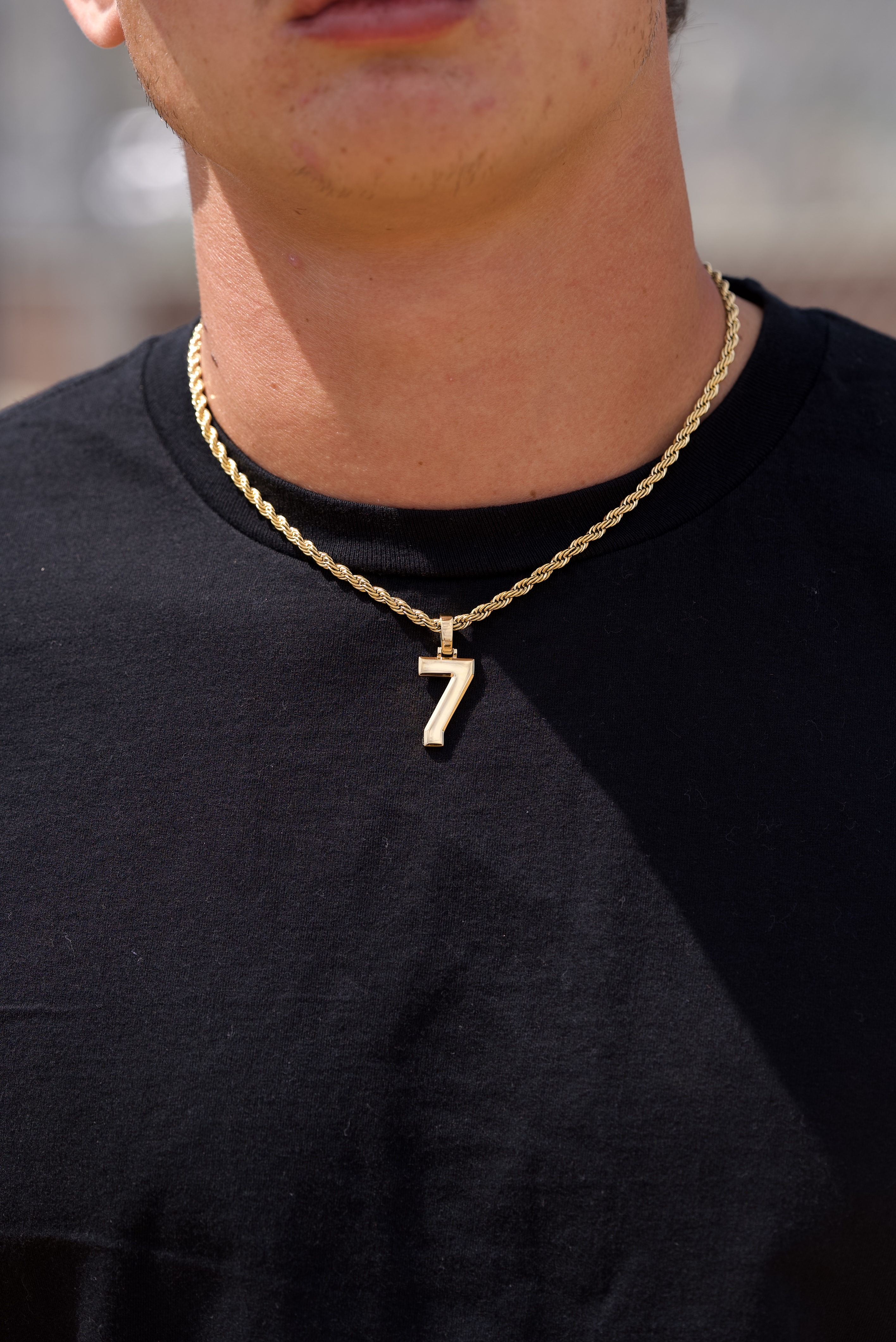 A close-up of a person wearing a black t-shirt and a gold rope chain with a number 7 pendant.