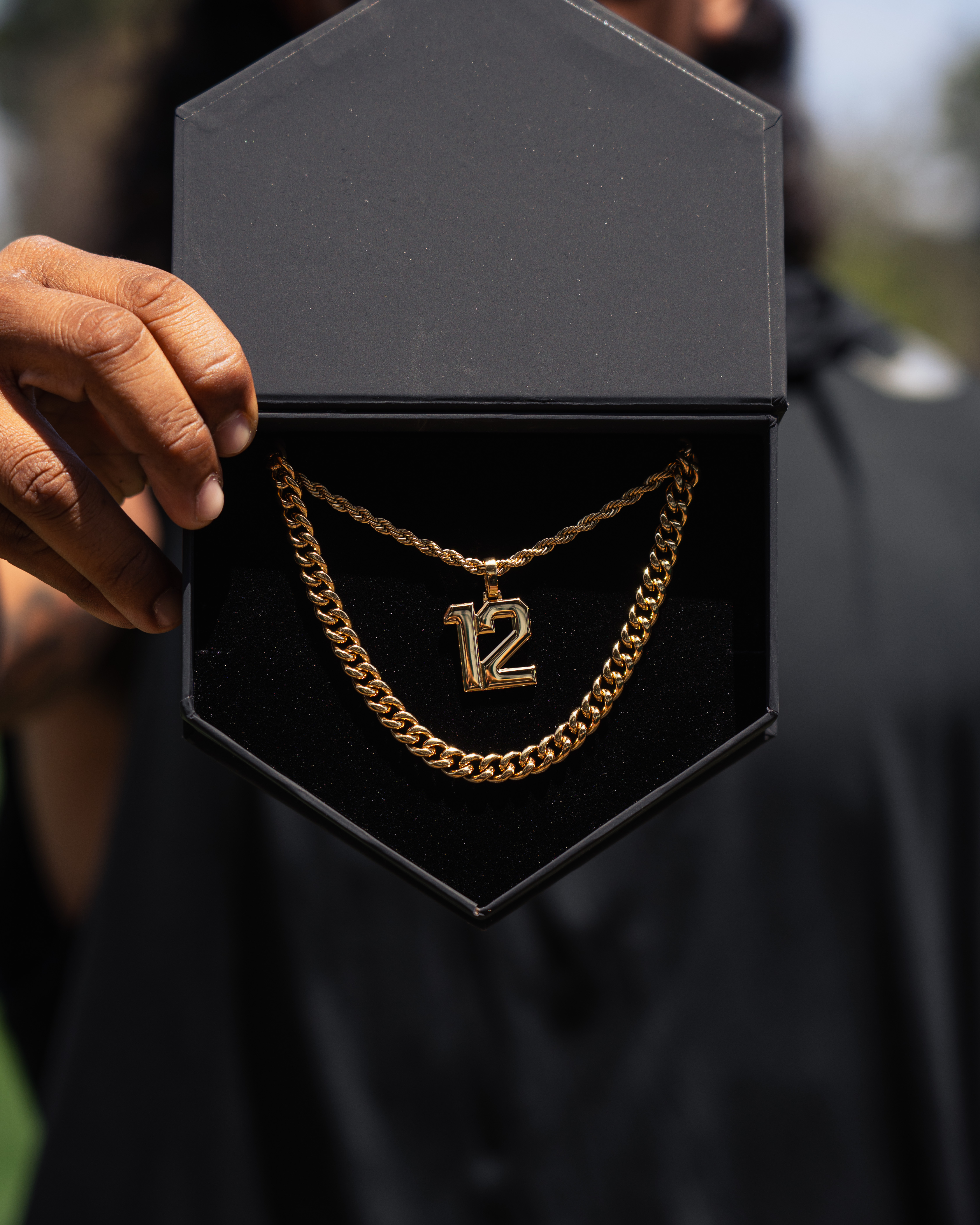 A person holds open a black box displaying a gold necklace with a number 12 pendant.