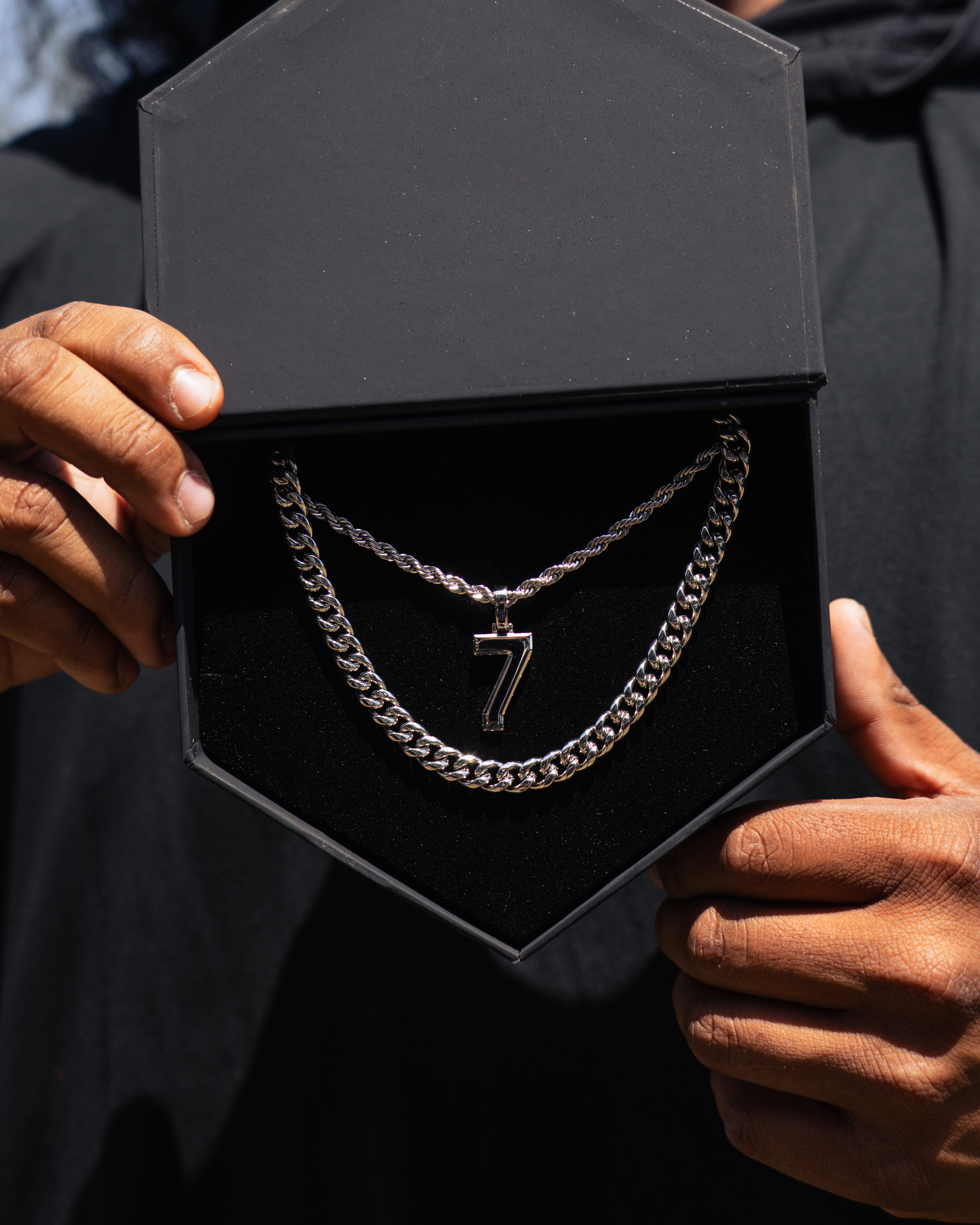 A person's hands hold open a black jewelry box revealing two silver chains and a number 7 pendant.
