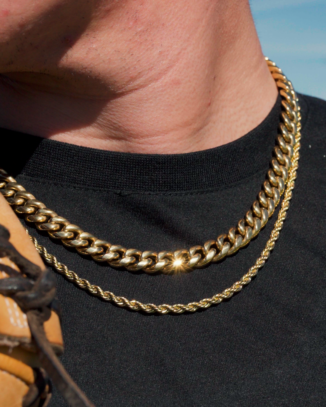 A close-up of a person's neck, showing two gold chains worn over a black t-shirt.