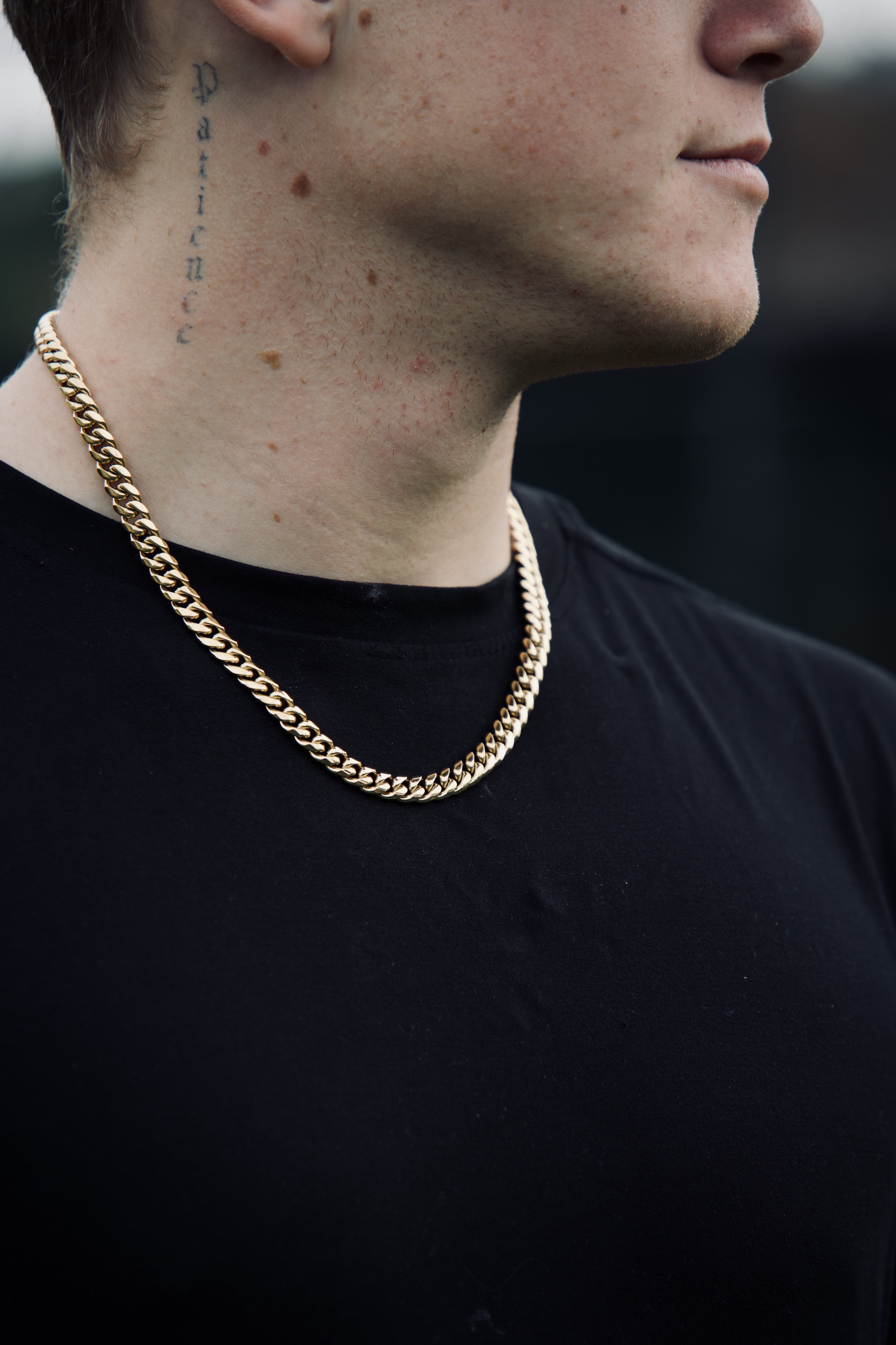A man with a neck tattoo wears a gold chain necklace and a plain black t-shirt.