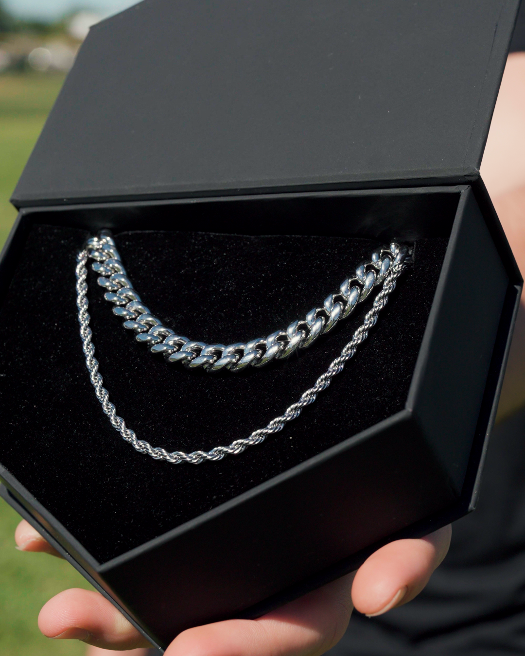 A hand holds open a black jewelry box containing two silver chains, a curb and a rope chain.