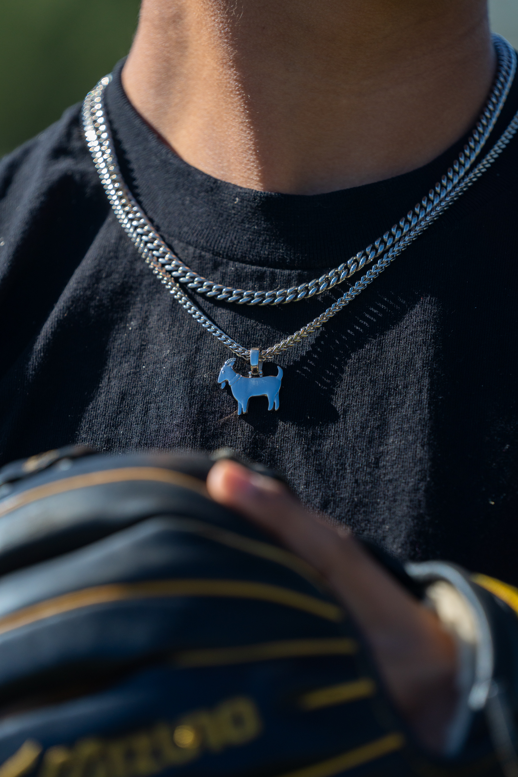 A close-up of a person wearing a silver necklace with a goat pendant, holding a baseball glove.