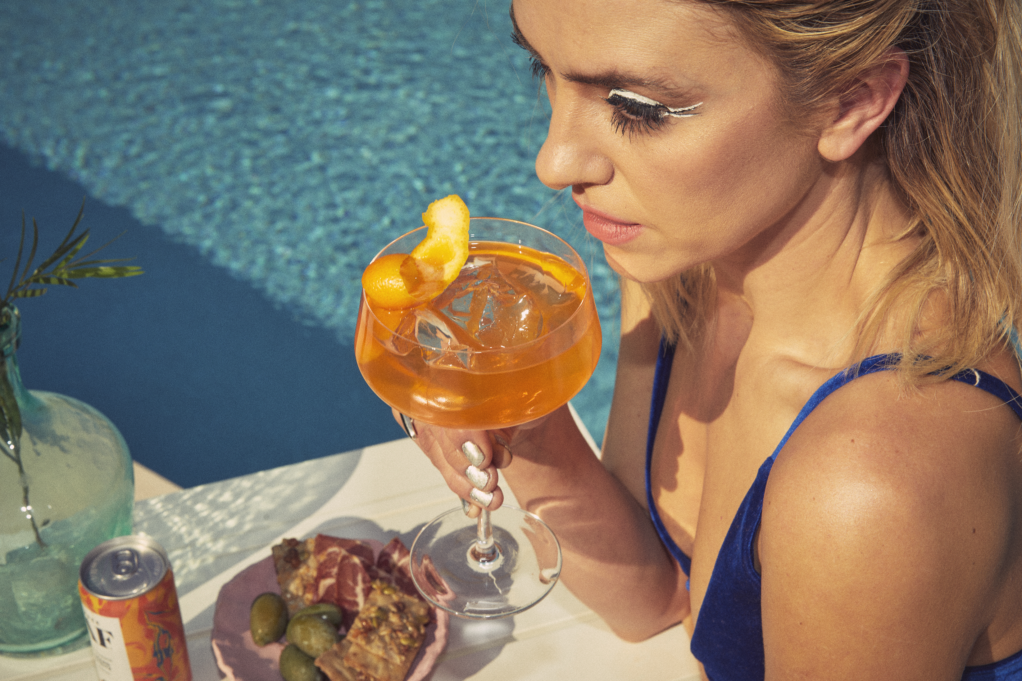 Woman holding a cocktail by the poolside with snacks on the table.