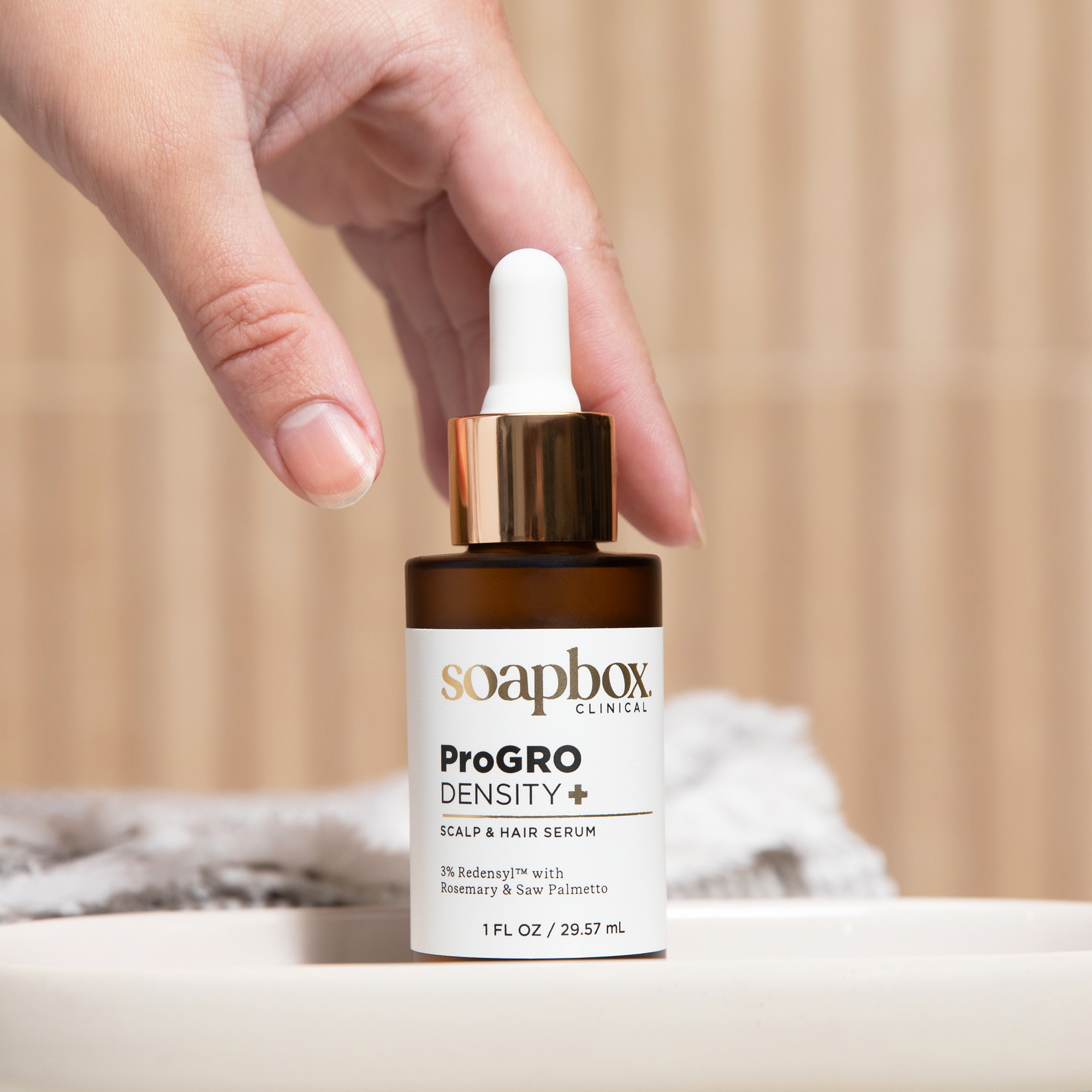 A hand holds a bottle of Soapbox Clinical ProGRO Density+ Scalp & Hair Serum over a sink.