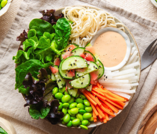 A bowl of noodles with vegetables, greens, and a small dish of sauce.