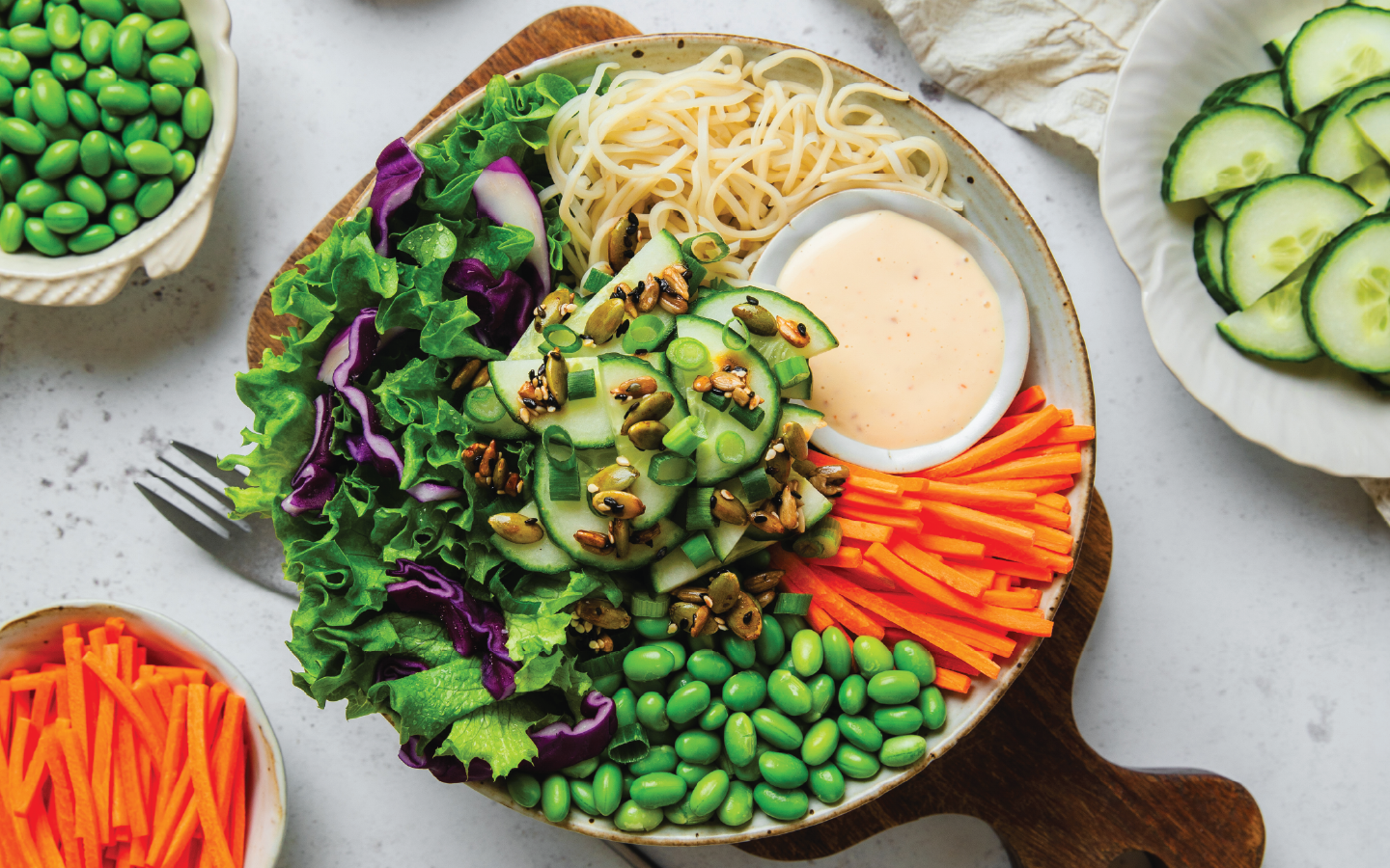 An overhead shot of a colorful salad bowl with edamame, carrots, cucumber, noodles, and a creamy dressing.