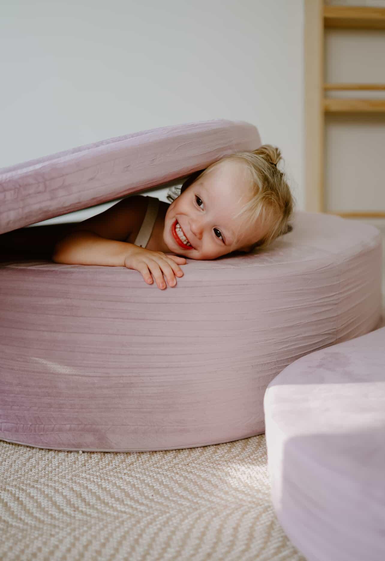 Child smiling, peeking out from between two large, soft cushions.