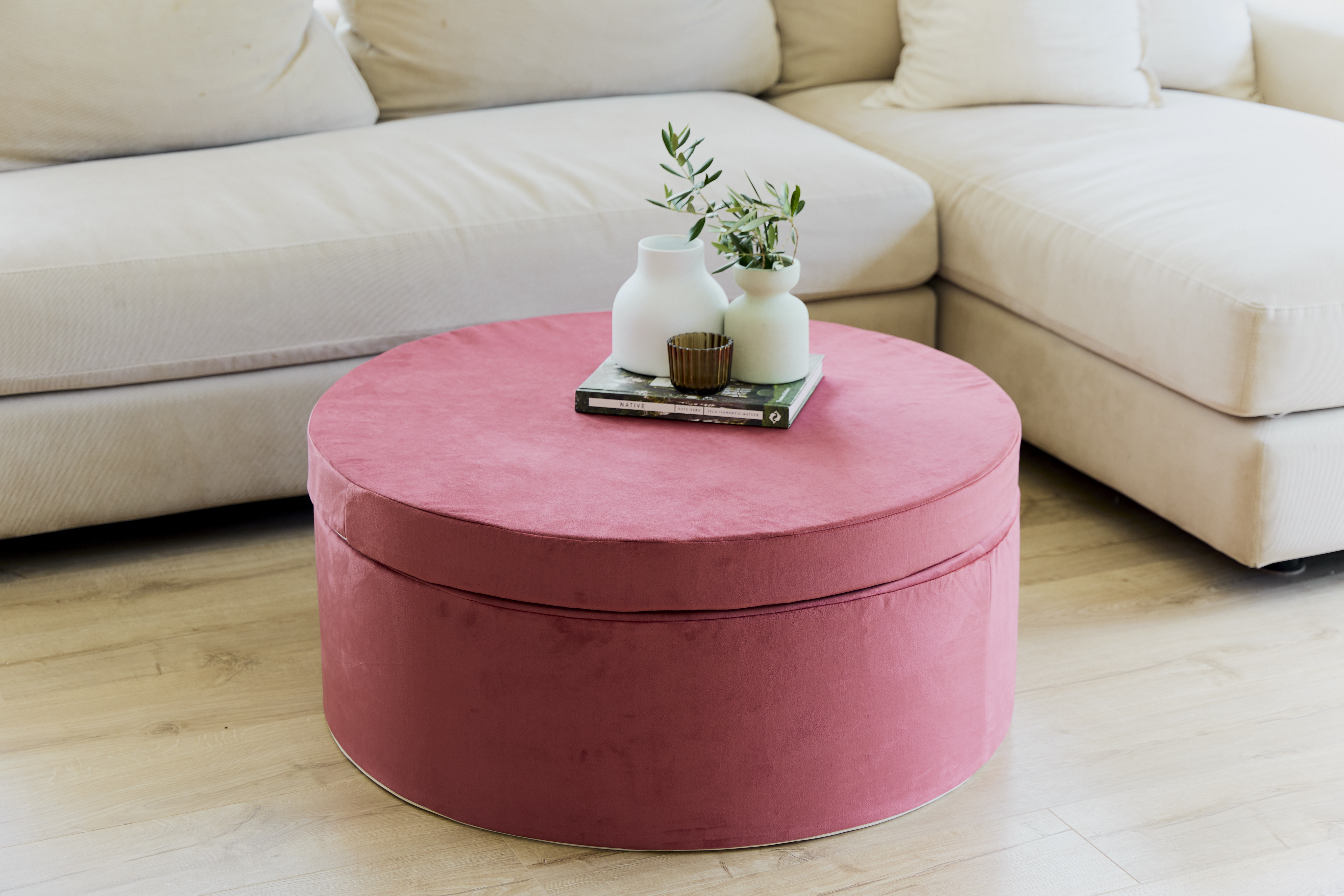 Round pink ottoman with vases and books, next to a beige sofa.