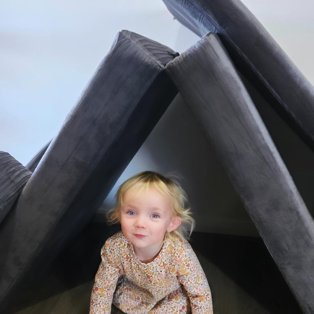 Child sitting under a makeshift tent made of gray cushions.