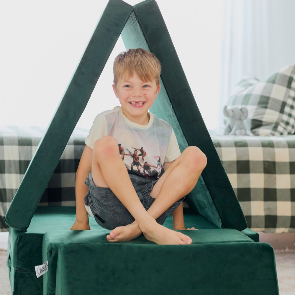 Child sitting inside a green triangular cushion fort, smiling.