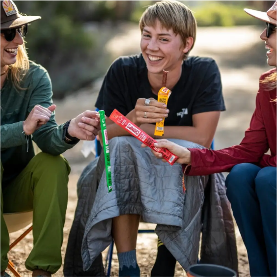 People sitting outdoors, smiling, and sharing snack sticks.