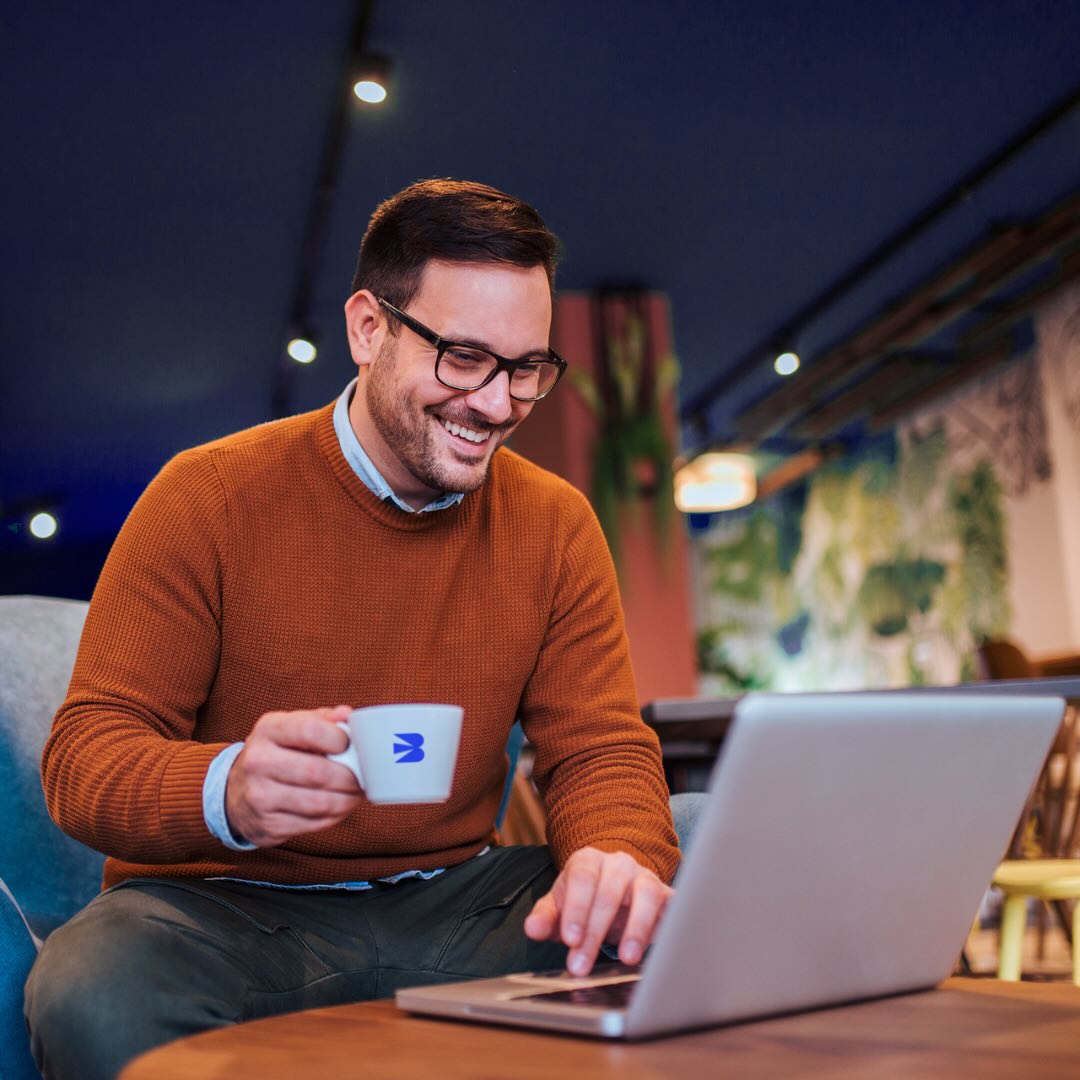 Man smiling while using a laptop and holding a coffee cup.