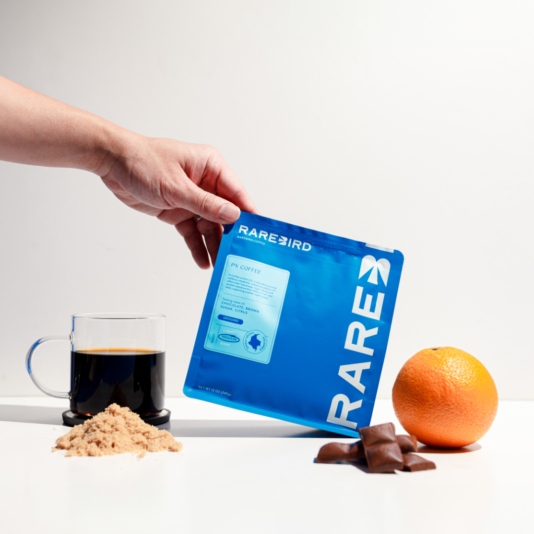 Hand holding blue coffee bag near coffee, orange, chocolate, and brown sugar.