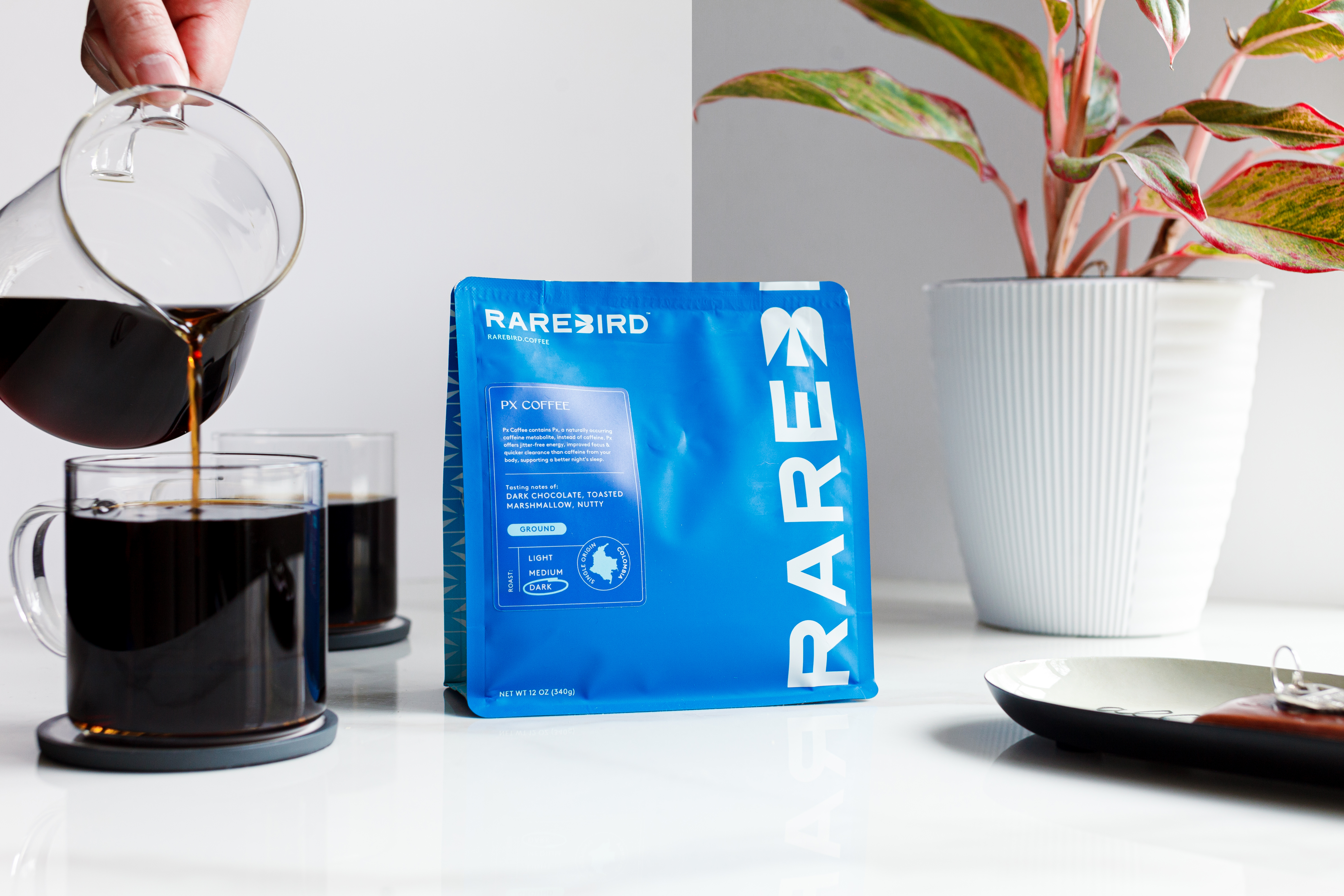 Pouring coffee into a glass mug beside a blue coffee bag and a potted plant.