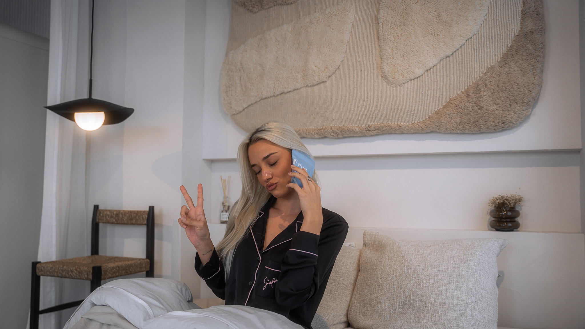 A woman in pajamas sits in bed, holding a blue pack to her face and making a peace sign.