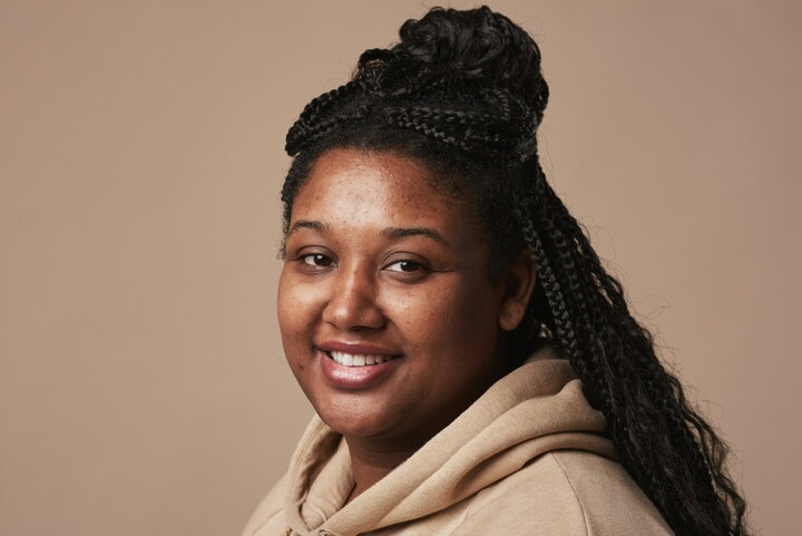 Smiling person with braided hair wearing a beige hoodie against a plain background.