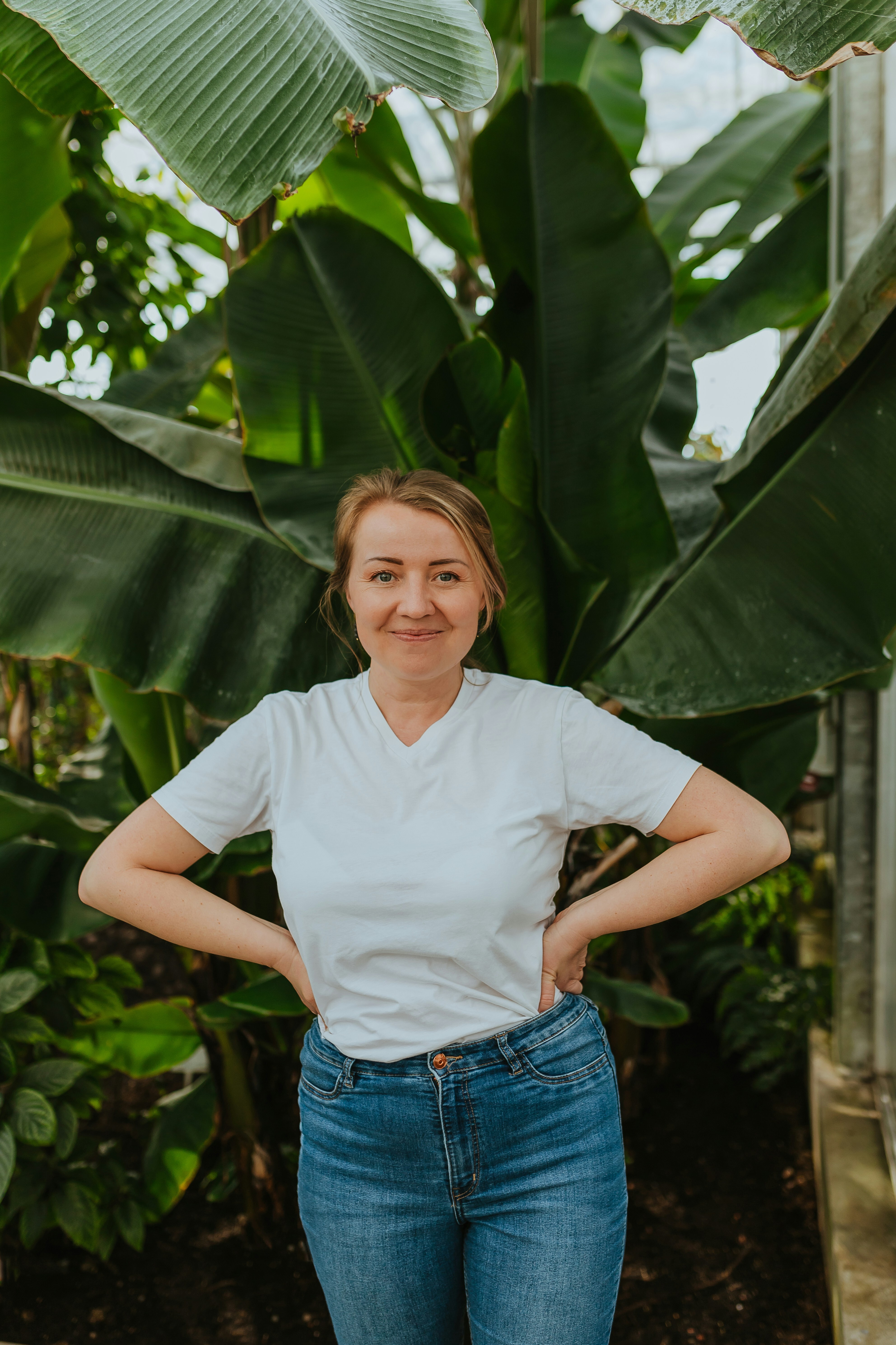 Person in white shirt and jeans standing in front of large green leaves.