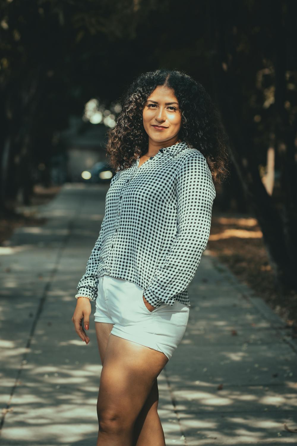 Woman in checked blouse standing on a sunlit sidewalk.