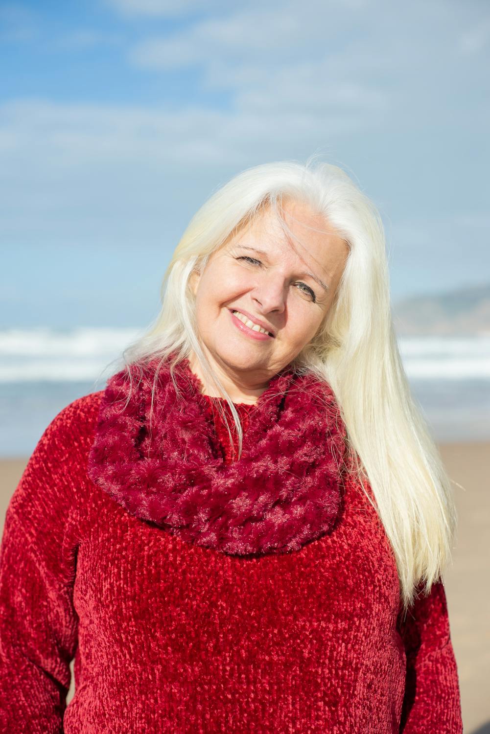 A woman with white hair wearing a red scarf and sweater on a beach.
