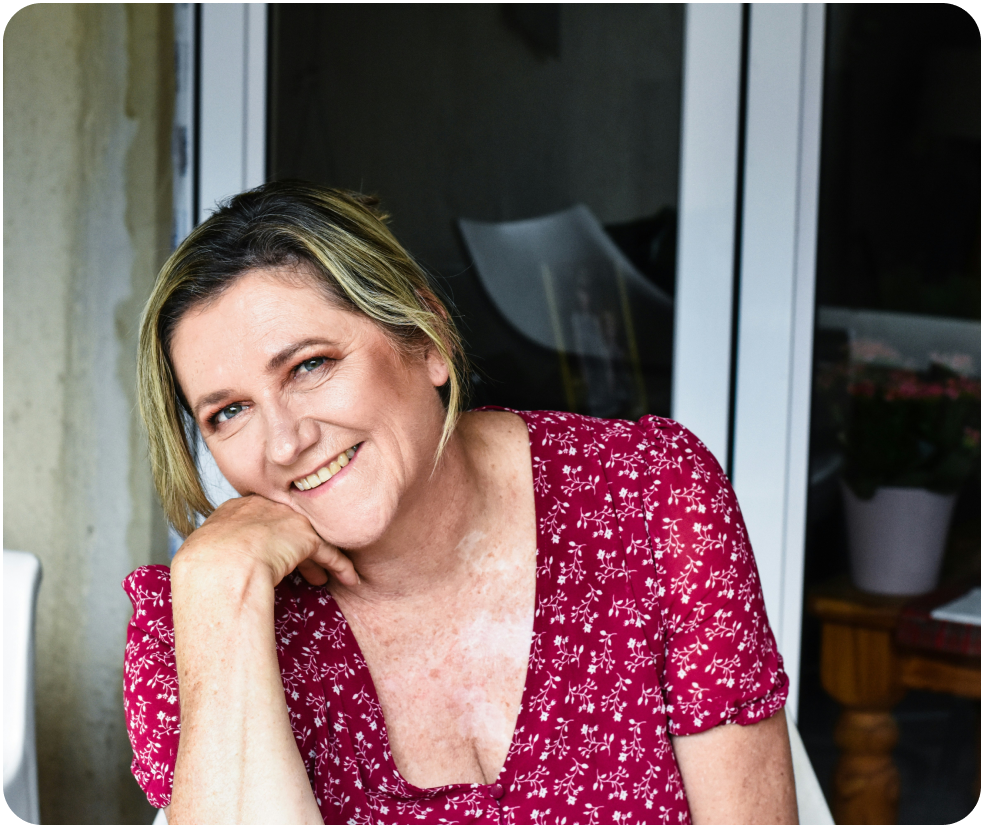 Smiling person in a red floral dress, sitting near a glass door.