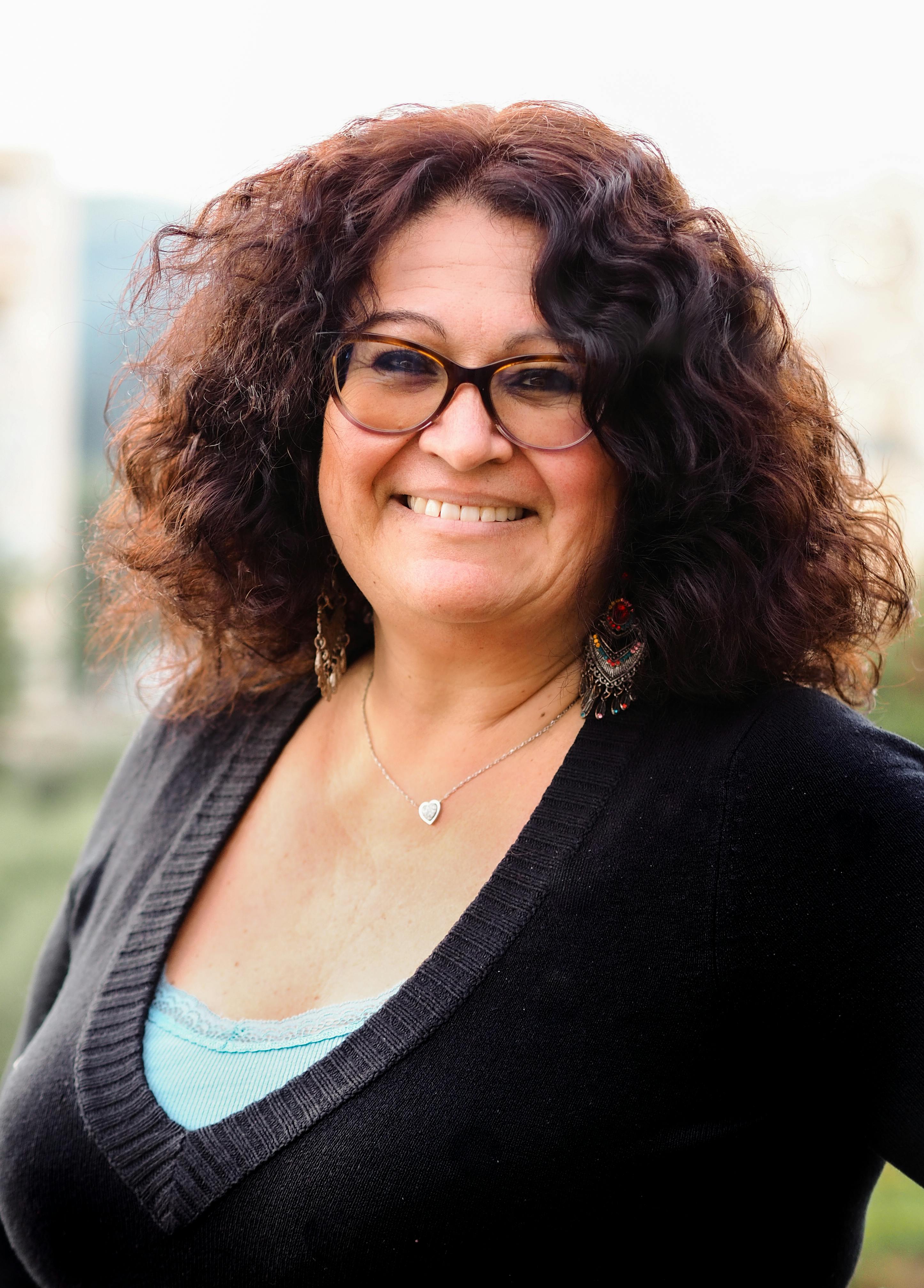 Smiling person with curly hair and glasses, wearing a black top with a heart necklace.