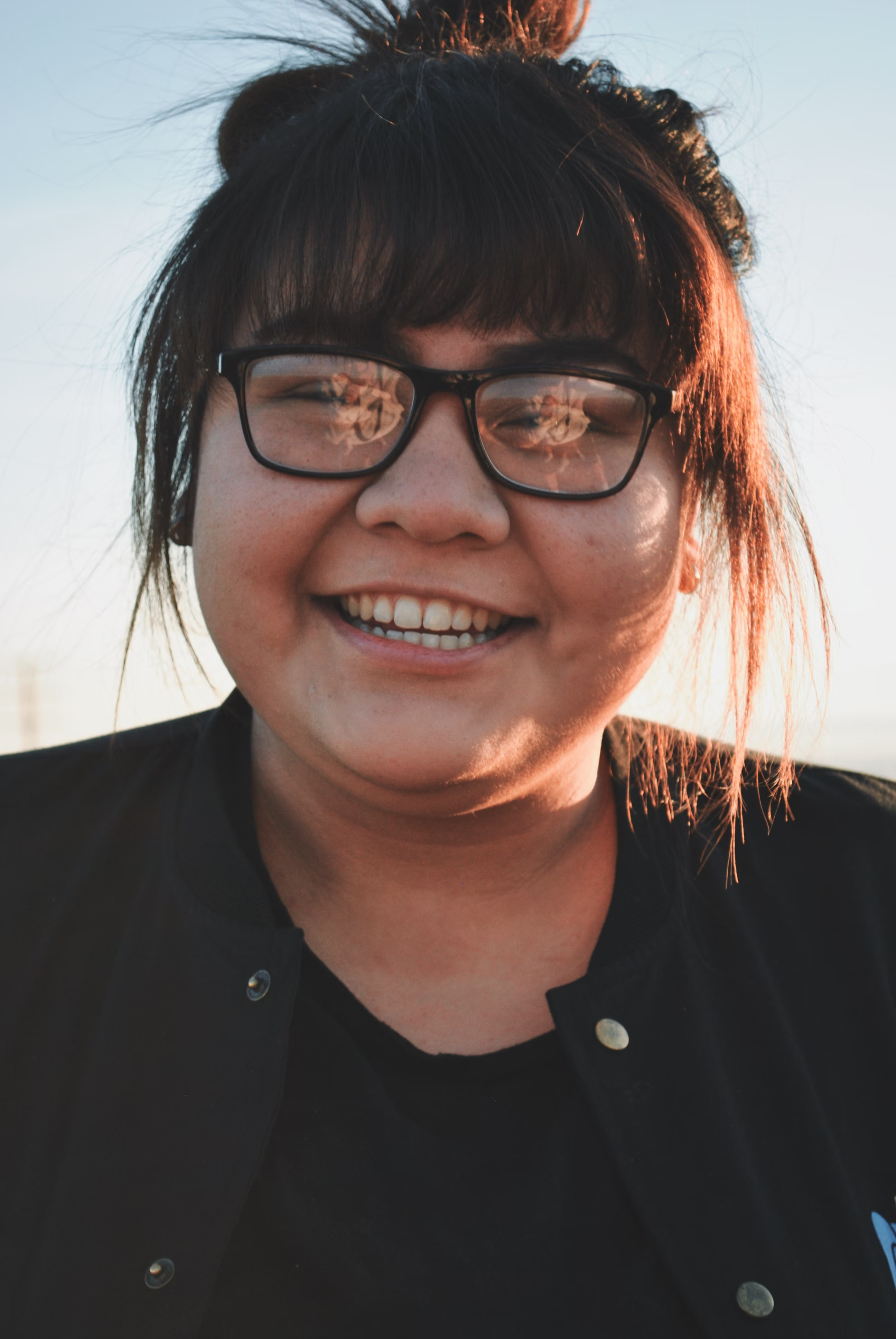 Smiling person with glasses and a bun, outdoors with sunlight.