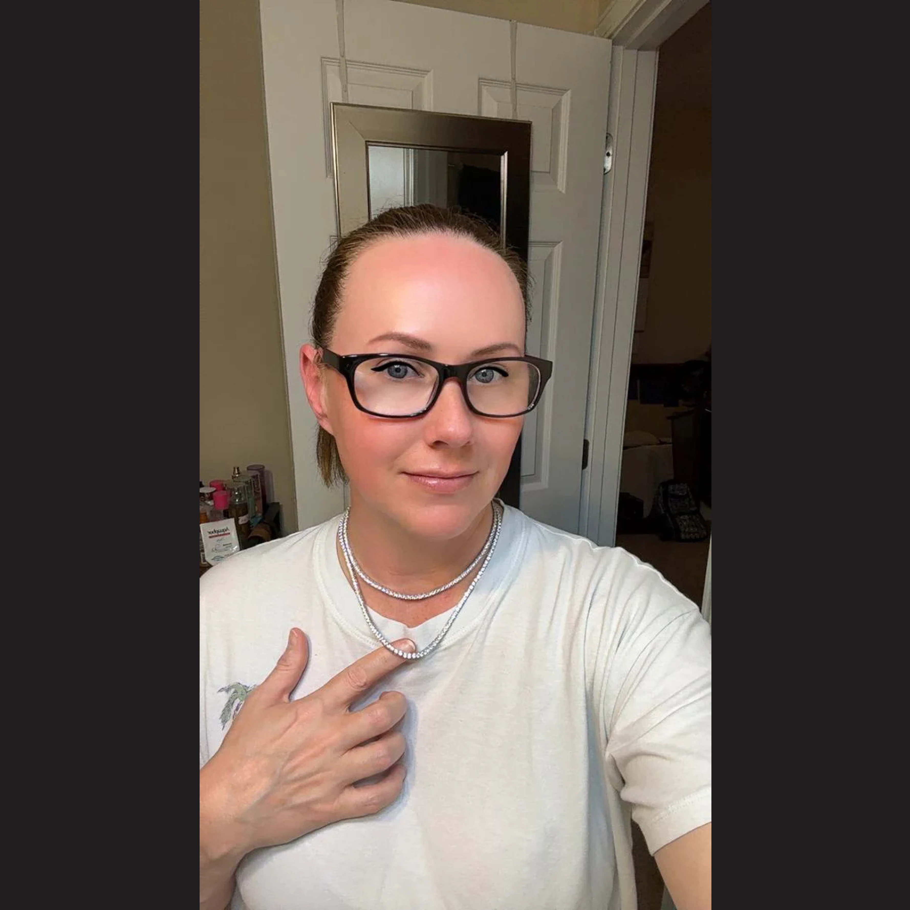 A woman wearing glasses and a white t-shirt points to her double-stranded necklace while taking a selfie.