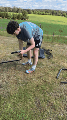 Person assembling metal frame outdoors on grass with fields in the background.