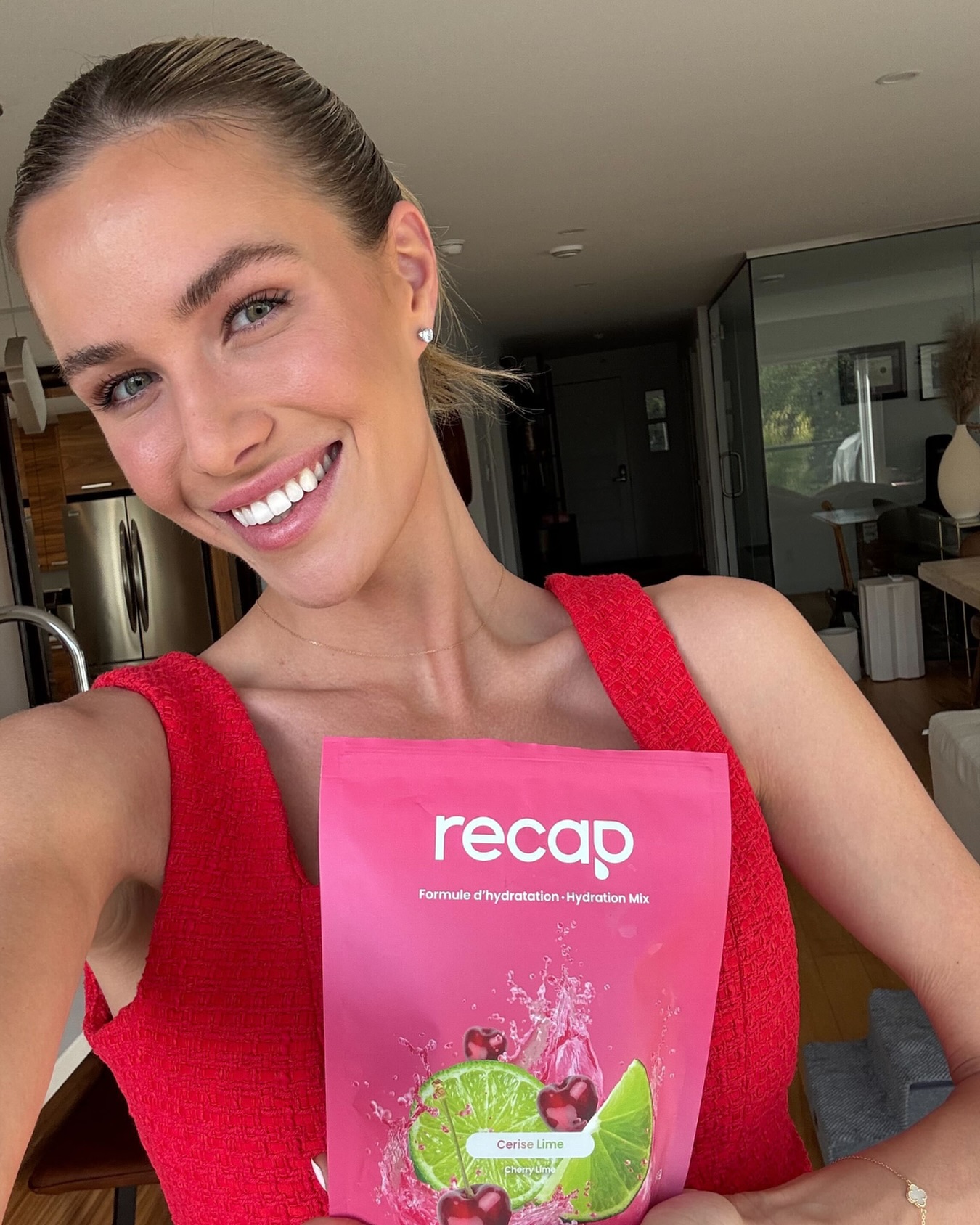 A smiling woman in a red top holds a pink pouch of Recap cherry lime hydration mix.