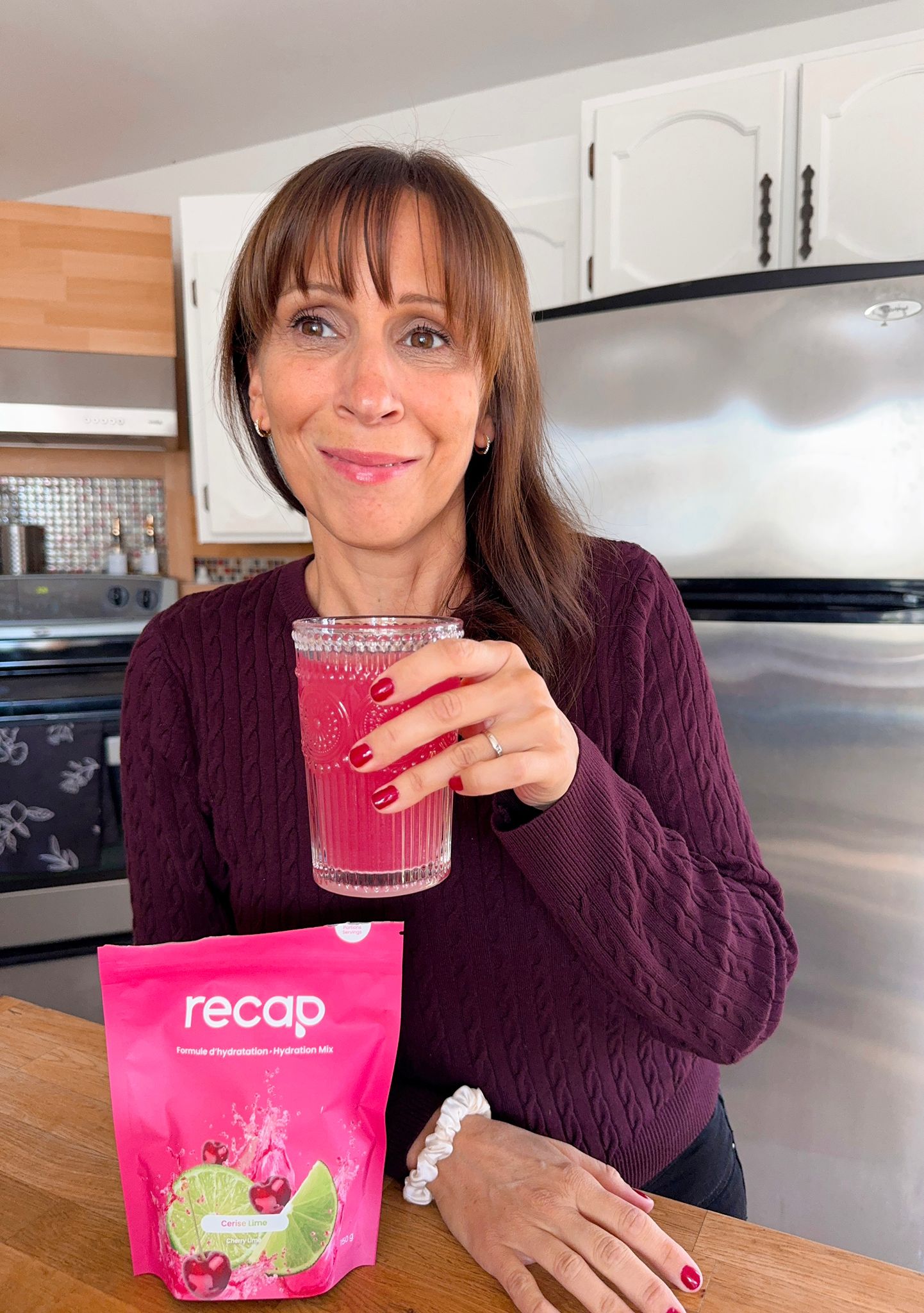 A woman holds a glass of pink drink, with a pouch of recap Hydration Mix on the table.