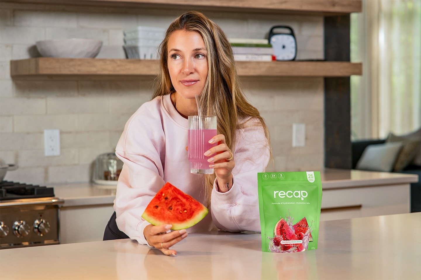 A woman in a kitchen holds a slice of watermelon and a pink drink next to a product pouch.