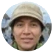 A close-up, circular photograph of a person wearing a tan cap and smiling.