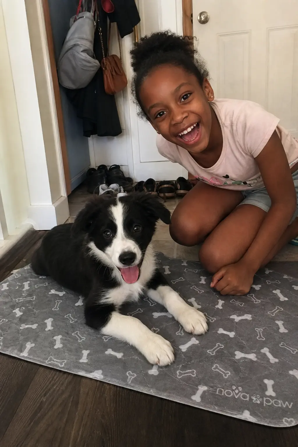 A young girl smiles widely while crouching next to a black and white puppy on the floor.