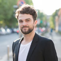 A man with dark curly hair and a beard, wearing a white shirt and black blazer, outdoors.