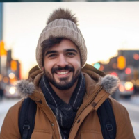A smiling man wearing a winter coat, scarf, and knit hat on a city street.