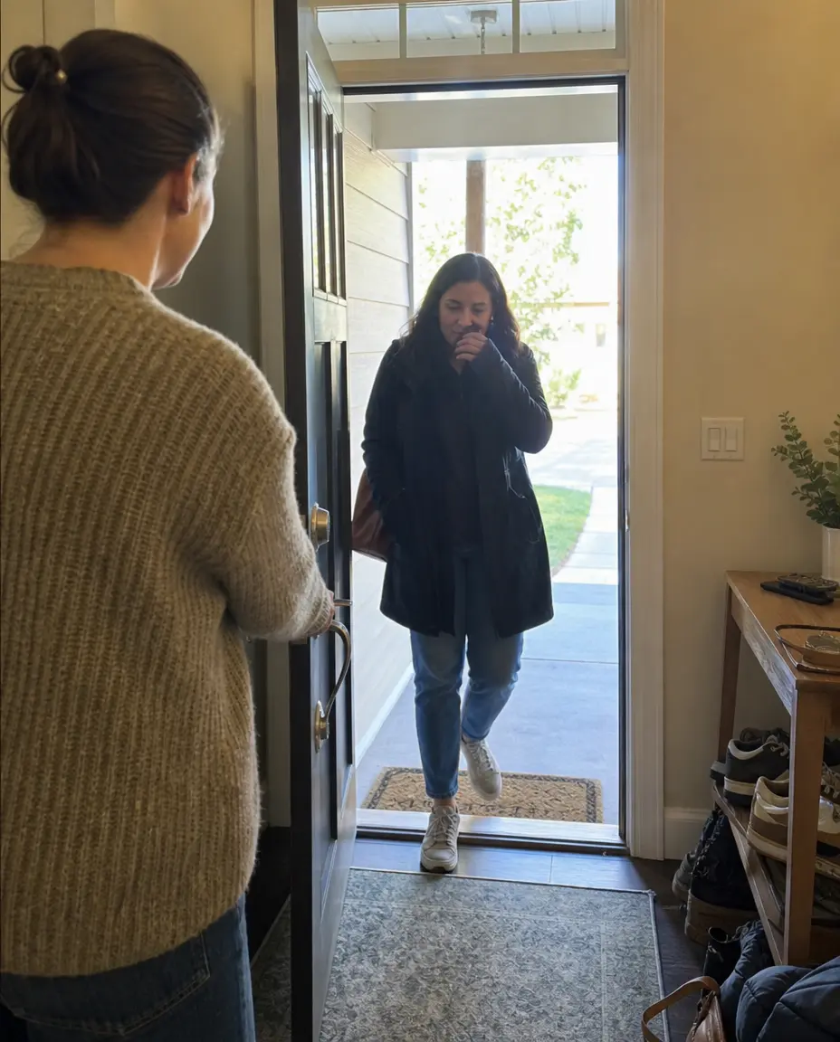 A woman holds a door open as another woman in a black coat walks into the house.