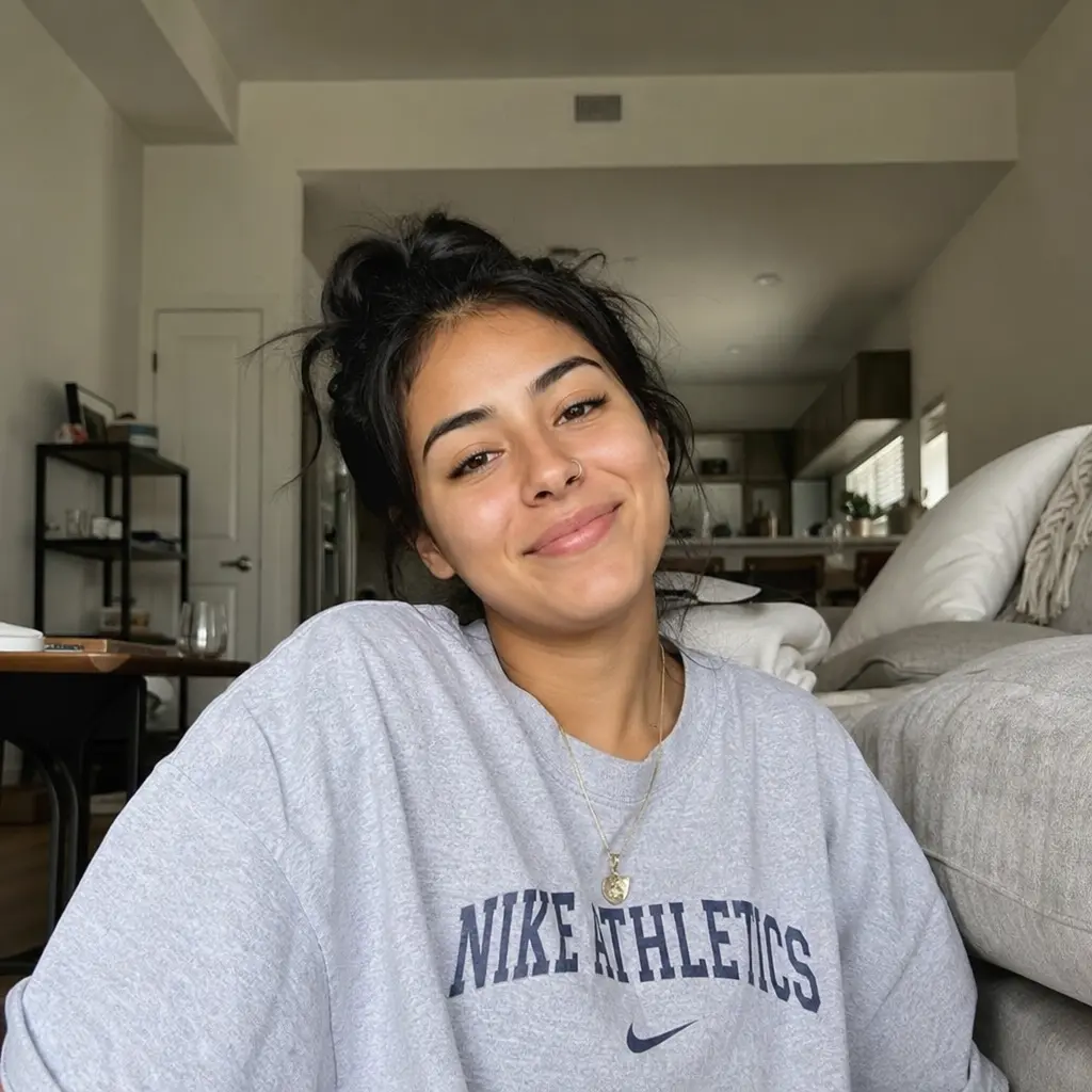 A smiling woman with dark hair in a messy bun wears a grey Nike Athletics sweatshirt indoors.