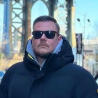 A man with a mustache wears sunglasses and a dark puffy coat outdoors with a bridge behind him.