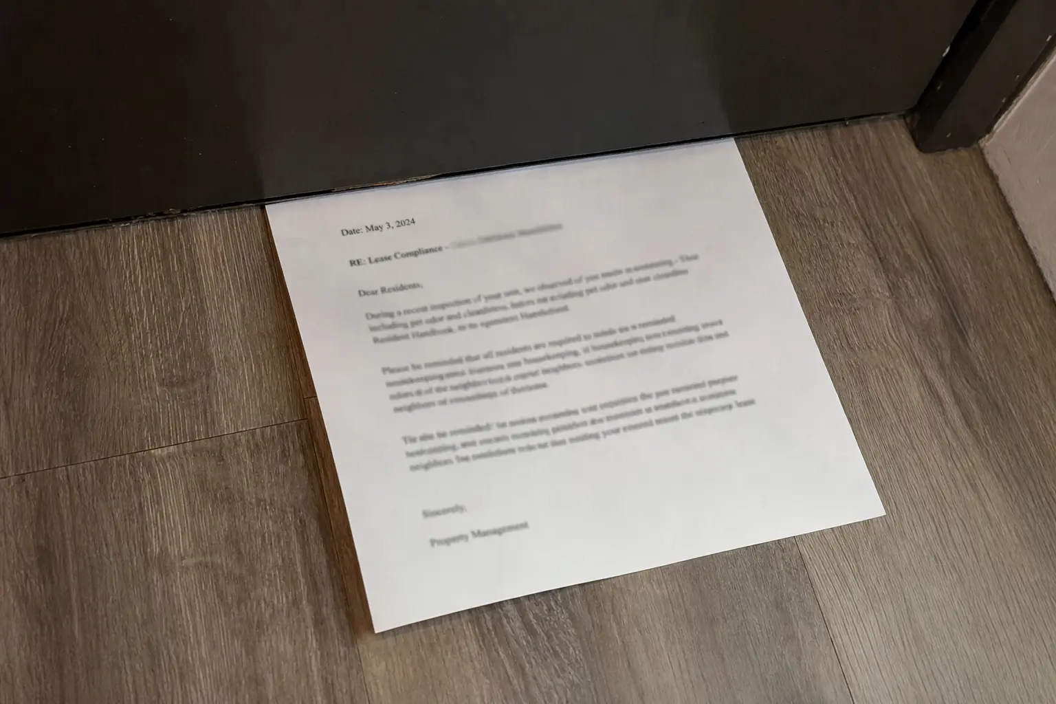 A lease compliance notice from property management slid under a door onto a wood-grain floor.