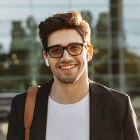 A smiling young man with dark hair and glasses, wearing wireless earbuds and a black blazer outdoors.