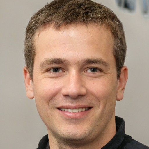 A headshot of a smiling man with short brown hair against a plain background.