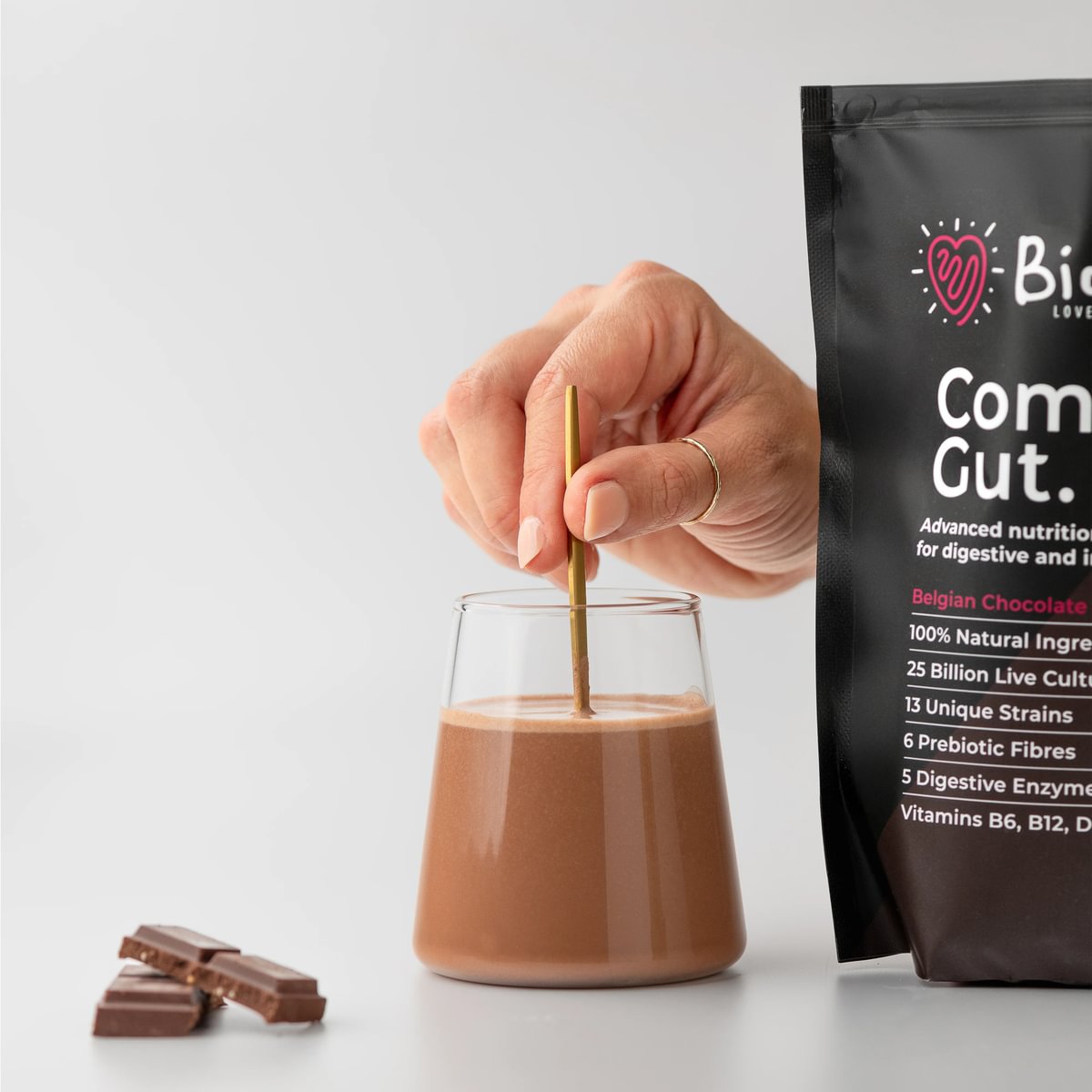 Hand stirring chocolate drink in a glass beside a product package and chocolate pieces.