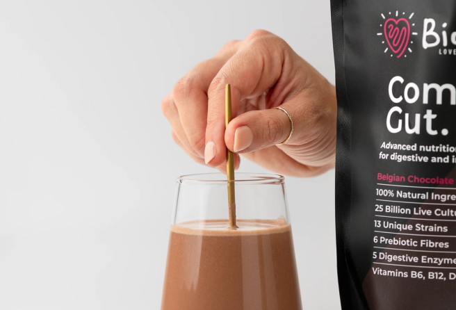 Hand stirring chocolate drink in a glass beside a nutritional supplement package.